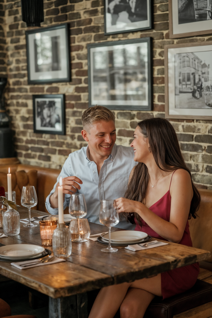 A happy couple is sitting at a cozy restaurant table, sharing a lighthearted moment and smiling at each other. The man, dressed in a light blue button-down shirt, and the woman, in a red dress, are engaged in cheerful conversation. The table is set with wine glasses, candles, and elegant place settings, creating a warm and intimate atmosphere. Behind them, the restaurant's brick wall is decorated with black-and-white framed photographs, adding to the vintage charm of the setting