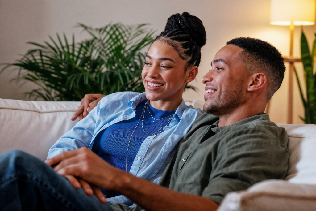 A happy couple sits close together on a comfortable white couch, smiling and enjoying a relaxed moment at home. The woman, with braided hair styled in a bun, is wearing a blue shirt and layered necklaces, resting her head on the man’s shoulder. The man, dressed in an olive green shirt, is leaning back casually. Behind them, lush green plants and a softly lit floor lamp create a cozy and inviting living room atmosphere. Both appear content and engaged in a peaceful moment together.