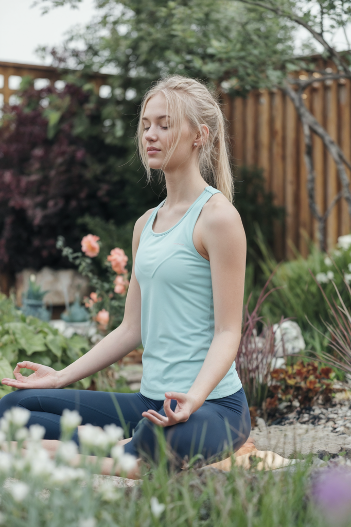 A young woman with blonde hair tied back in a ponytail is sitting cross-legged in a peaceful garden, practicing meditation. She wears a light blue athletic tank top and navy blue leggings, with her eyes closed and hands resting on her knees in a relaxed, meditative posture. The surrounding garden is lush with greenery and blooming flowers, creating a serene and natural environment. The wooden fence and trees in the background add to the tranquil outdoor setting