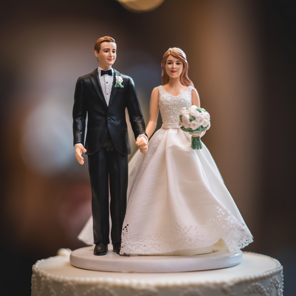 A close-up of a bride and groom cake topper placed on a wedding cake. The figurines depict the groom in a classic black tuxedo and bow tie, and the bride in a white gown with lace detailing, holding a bouquet of flowers. They are standing hand in hand, symbolizing their union. The cake beneath the topper is elegantly decorated with white frosting, while the soft lighting in the background creates a warm and romantic atmosphere, highlighting the details of the topper.