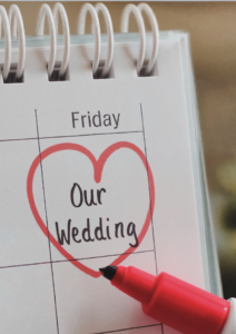 A close-up of a calendar with a red marker drawing a heart around the words "Our Wedding" written on a Friday. The red marker is visible in the corner, emphasizing the excitement and importance of the wedding day. The calendar is bound with a spiral ring, and the image captures the moment of planning or marking the special day. The heart and bold writing symbolize love and the anticipation of the upcoming celebration.