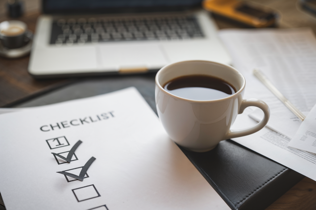 A wedding planning checklist on a desk with two items checked off, a cup of coffee, and a laptop in the background, symbolizing organized wedding preparation.