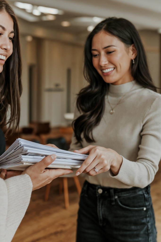 Smiling woman handing over a stack of papers, wearing an engagement ring and casual outfit, engaging in a friendly exchange.