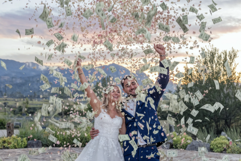 Happy bride and groom celebrating outdoors with flower petals and dollar bills falling from the sky, creating a festive and joyful wedding scene against a scenic mountain backdrop.