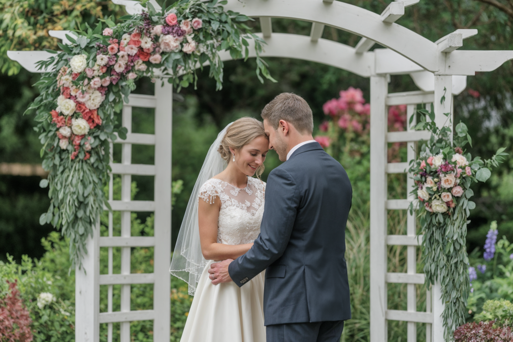 Bride and groom share an intimate moment under a floral arch at their outdoor garden wedding ceremony.